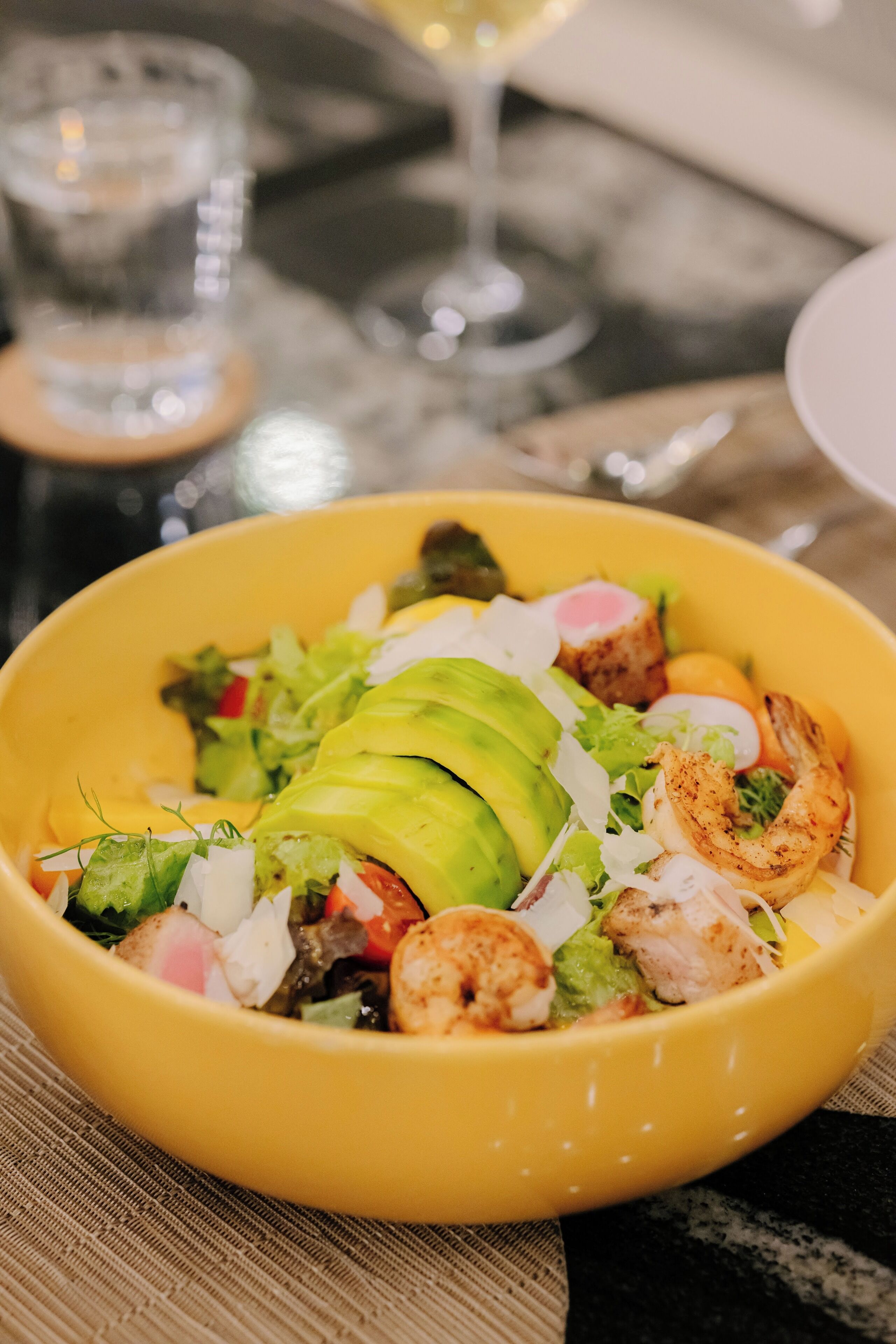 Uma saladeira amarela com salada de abacate, camarão e legumes sobre uma mesa.