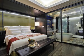 Quarto de hotel moderno com uma cama grande, banheiro com paredes de vidro e vista para a paisagem urbana.