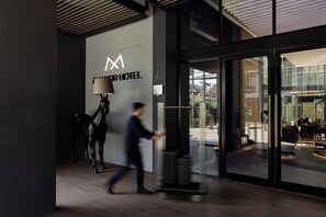 Entrada de hotel moderna, com um grande logotipo, um balcão de recepção e uma porta de vidro que dá acesso ao interior.