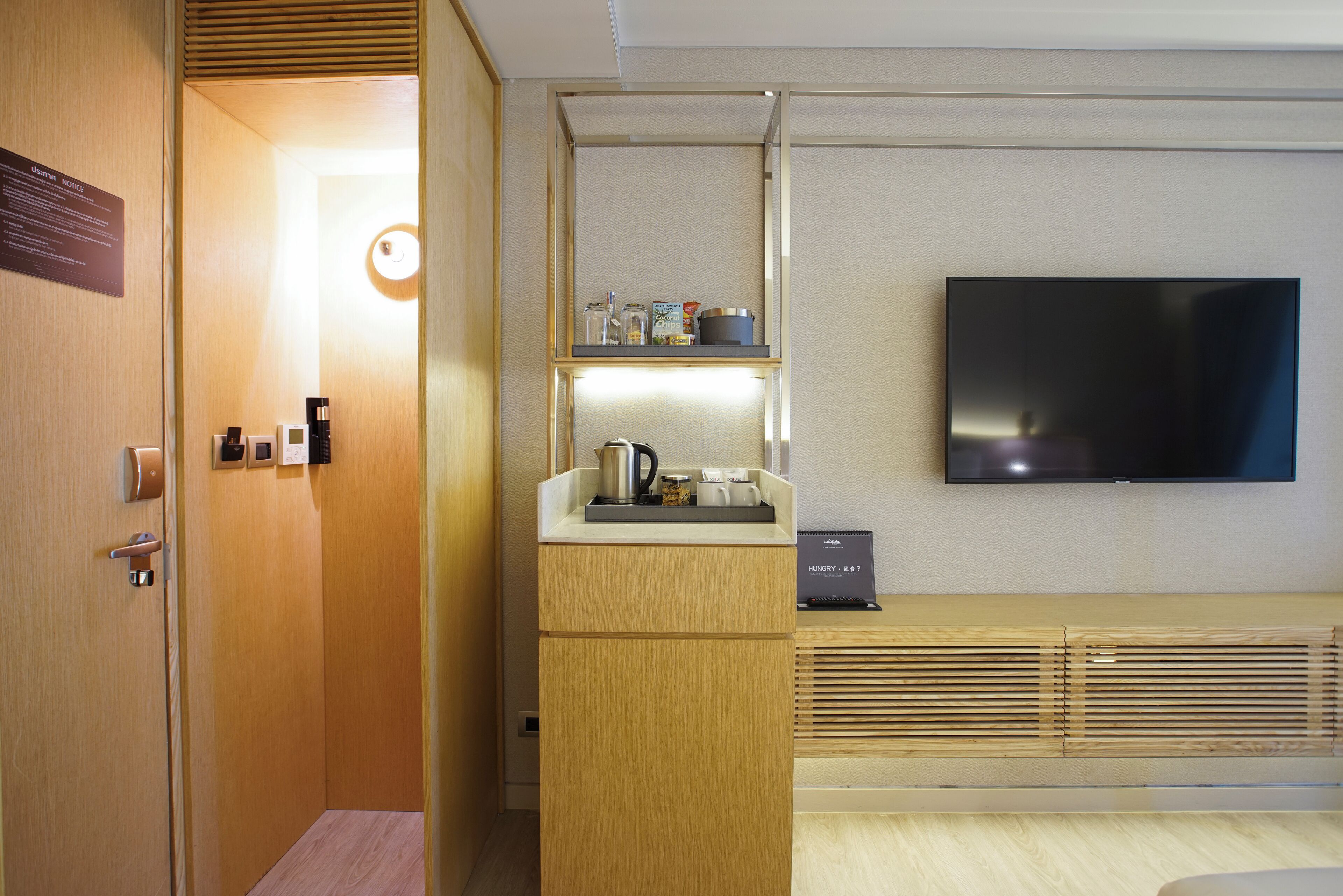 Ein Hotelzimmer mit Holztür, einer kleinen Küchenzeile mit Kaffeemaschine, einem Flachbildfernseher und einem Schild mit der Aufschrift 'HUNGRY'.