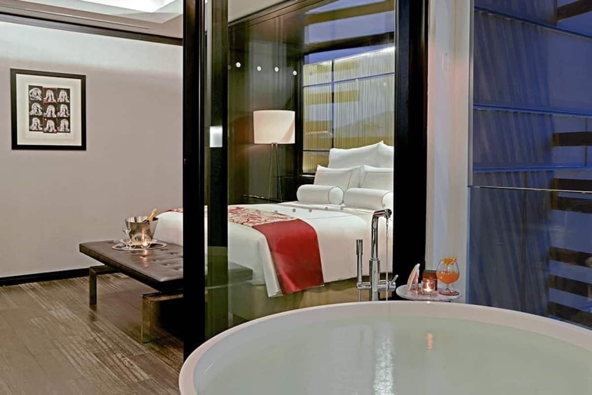Ein modernes Hotelzimmer mit einer großen Badewanne, einem Bett mit weißer Bettwäsche, einem Nachttisch mit Lampe und einem gerahmten Bild an der Wand.