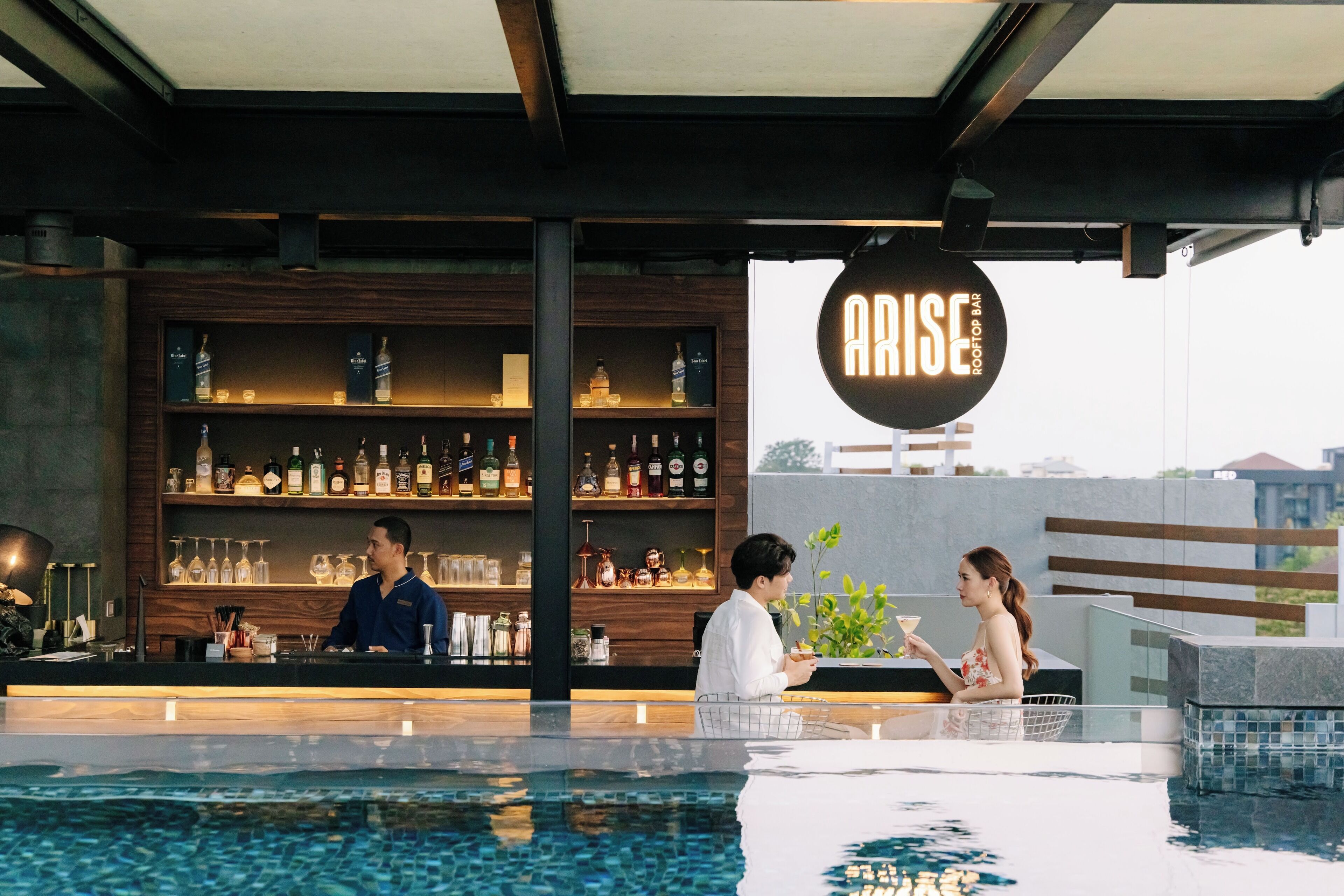 A rooftop bar with a pool, bar shelves stocked with bottles, and a sign reading "ARISE ROOF BAR".
