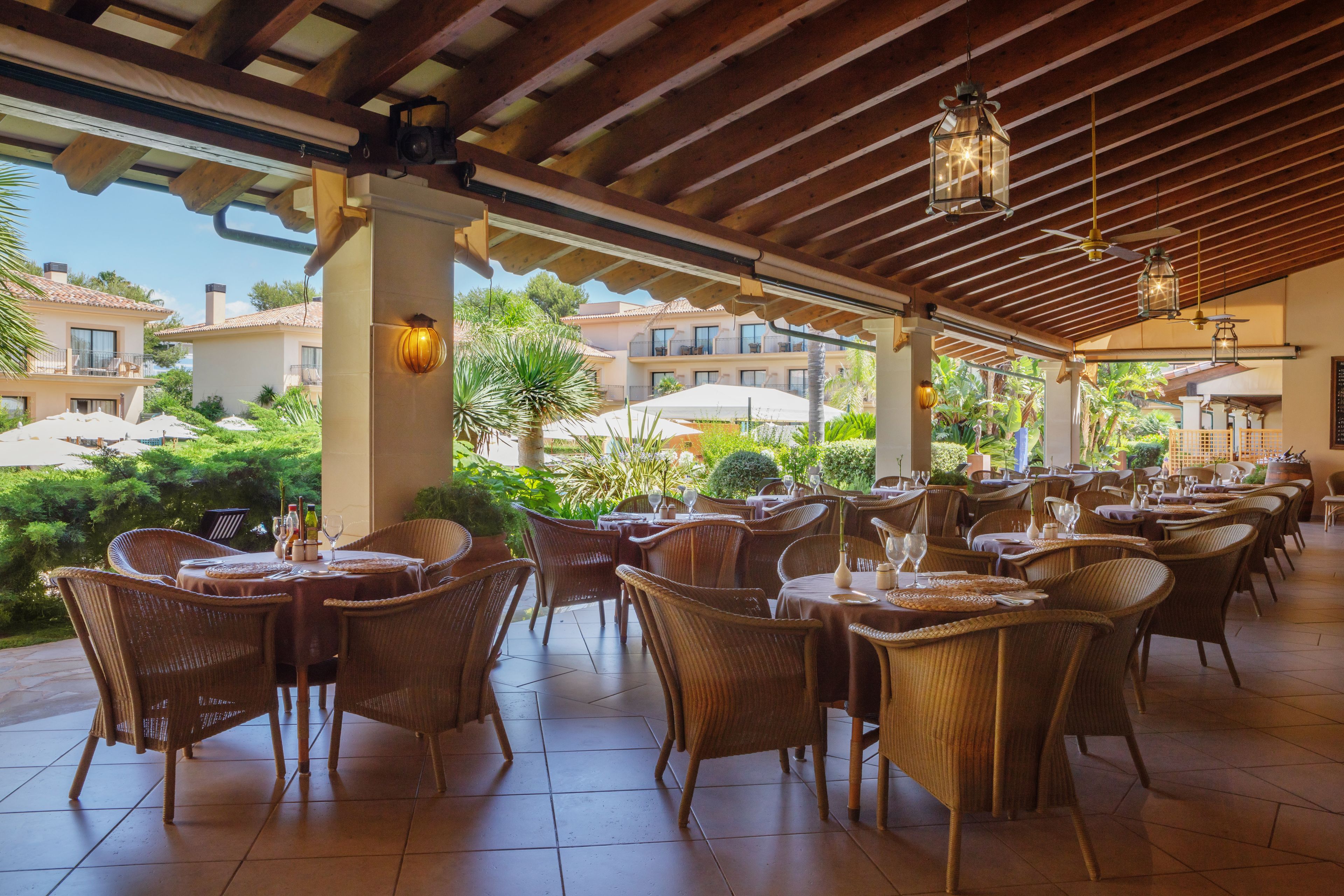 An outdoor patio with wicker furniture and umbrellas, overlooking a landscaped garden and buildings.