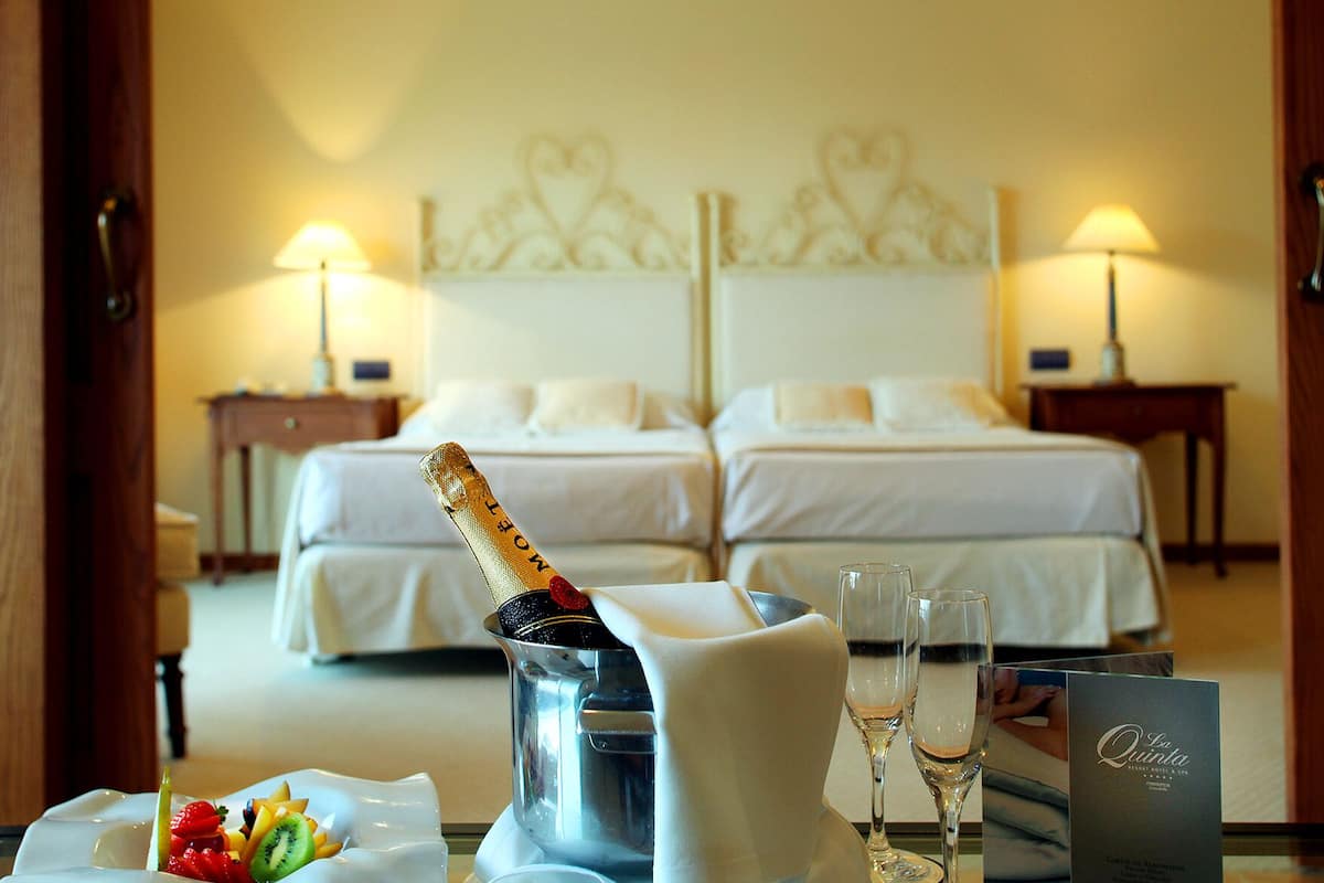Quarto de hotel com duas camas, minibar e uma mesa com champanhe, copos e frutas.
