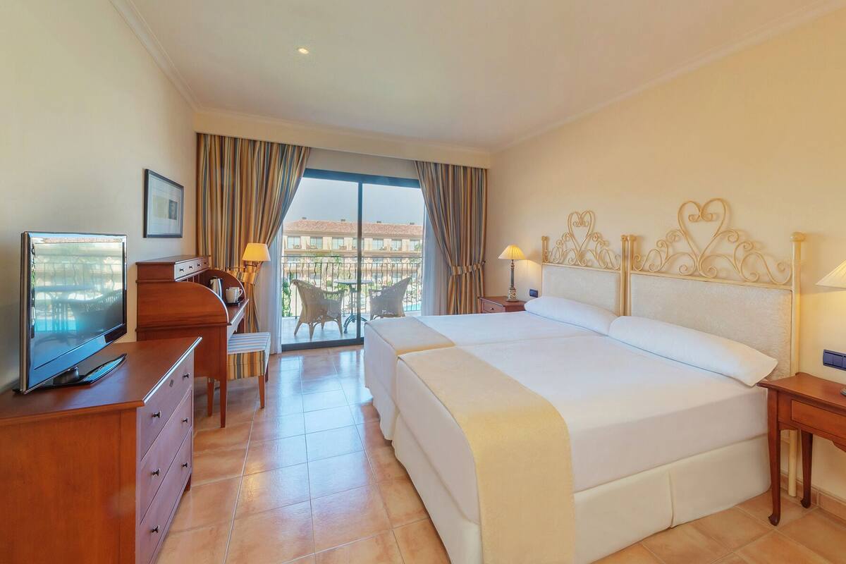Quarto de hotel com uma cama grande, televisão, escrivaninha e varanda com mobiliário para área externa.