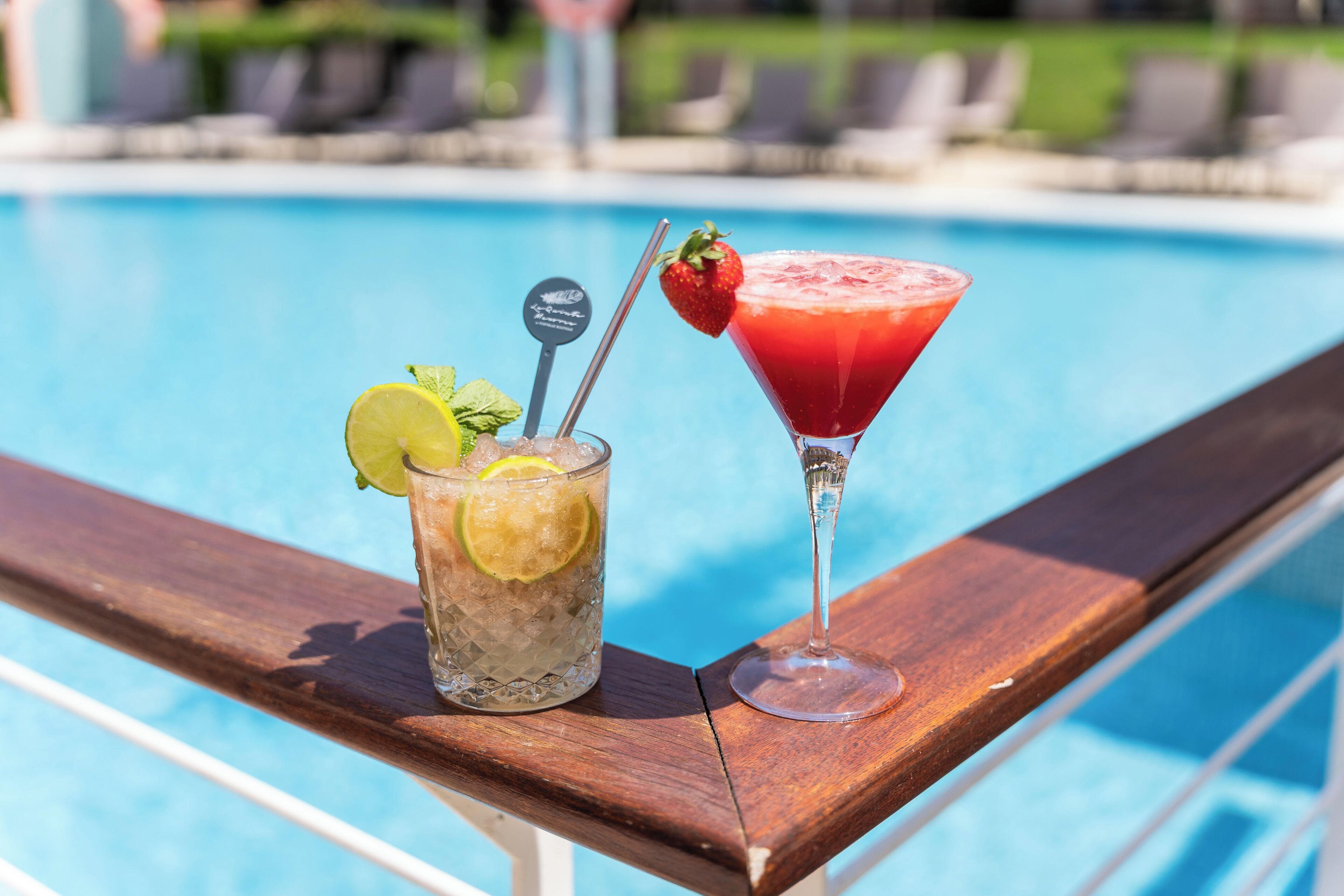 Dos bebidas sobre una mesa de madera al lado de la piscina, con una piscina al fondo.