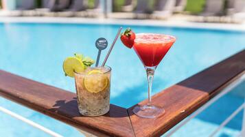 Dos bebidas sobre una mesa de madera al lado de la piscina, con una piscina al fondo.