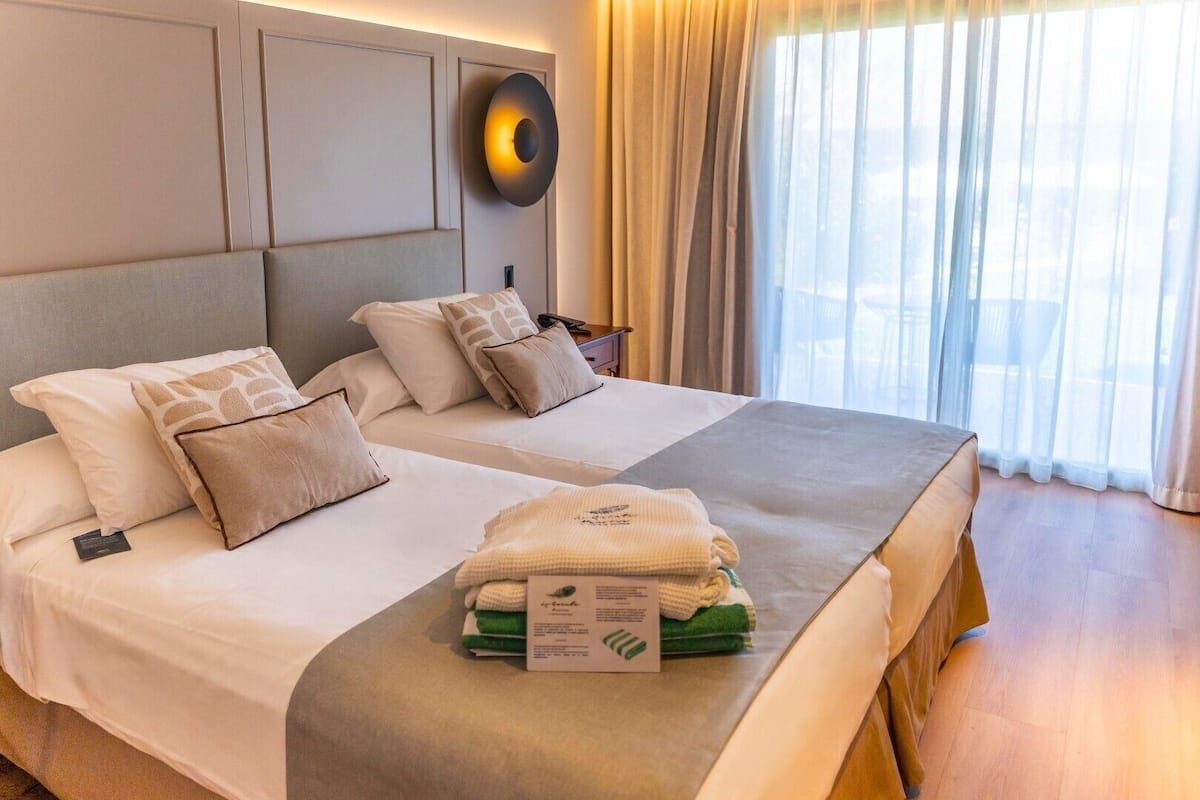 Quarto de hotel com duas camas, uma janela grande com cortinas, luminária fixada na parede e cama bem arrumada com toalhas e um cartão de boas-vindas.