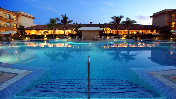 A large, clear swimming pool with a central pole, surrounded by palm trees and a building with multiple balconies. - La Quinta Menorca by Portblue Boutique (Ciutadella de Menorca)