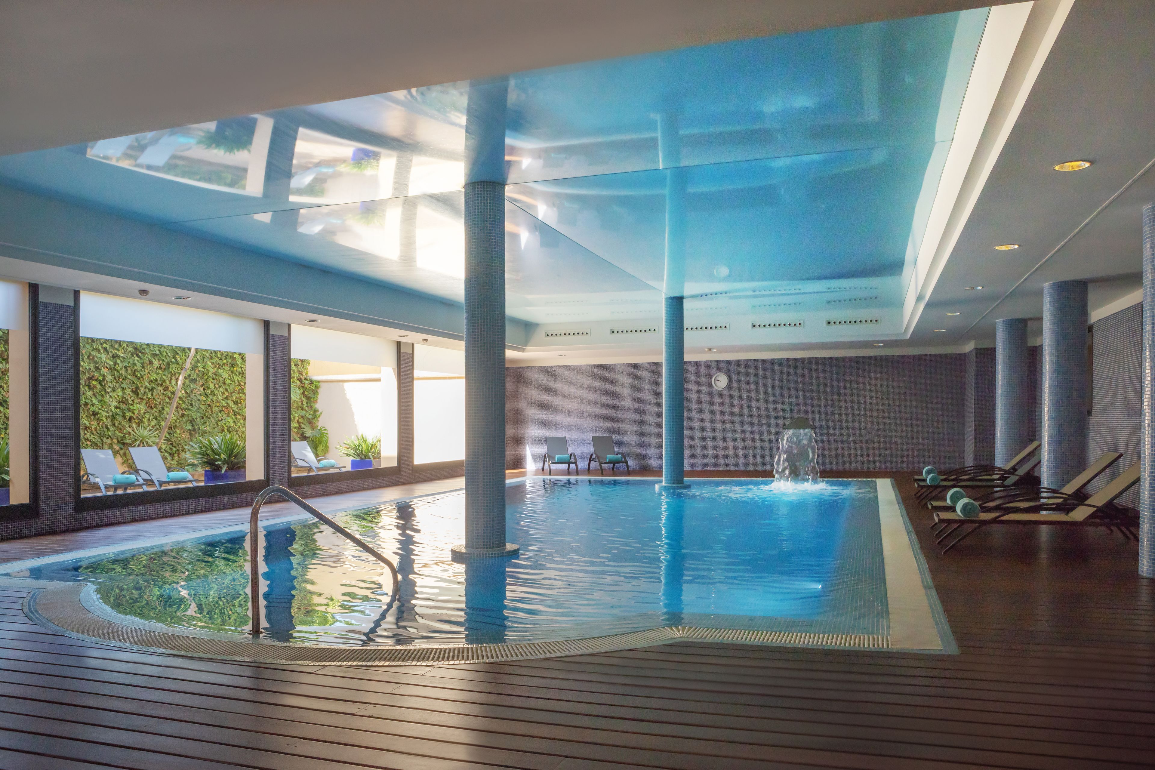 An indoor swimming pool with clear water, lounge chairs, and a view of greenery outside.