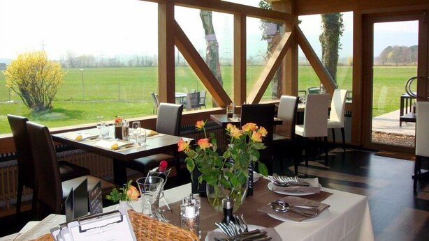 Een eetruimte met tafels voor twee personen, grote ramen met uitzicht op een groen veld, en houten balken in het plafond.