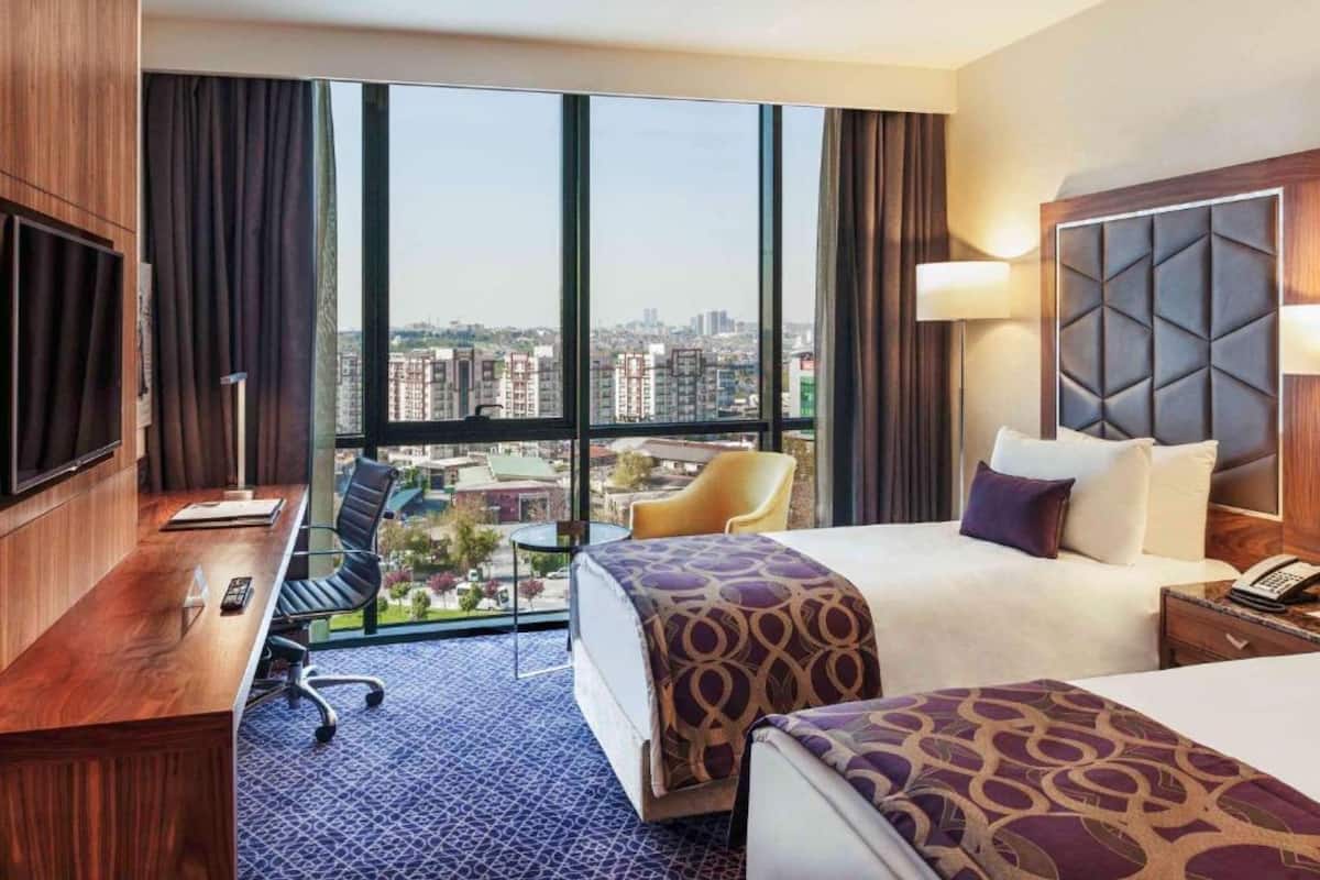 Habitació d'hotel amb una finestra gran, un llit, un escriptori amb una cadira i una televisió.