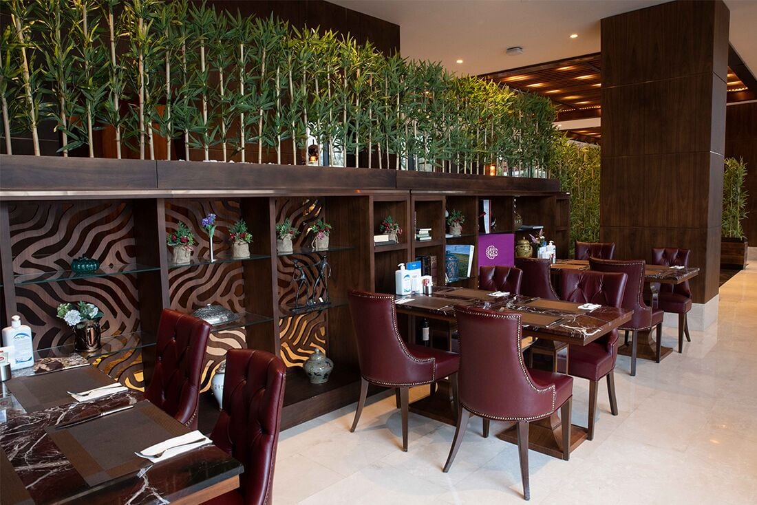A restaurant with a long counter, red upholstered chairs, and a wall of potted plants.
