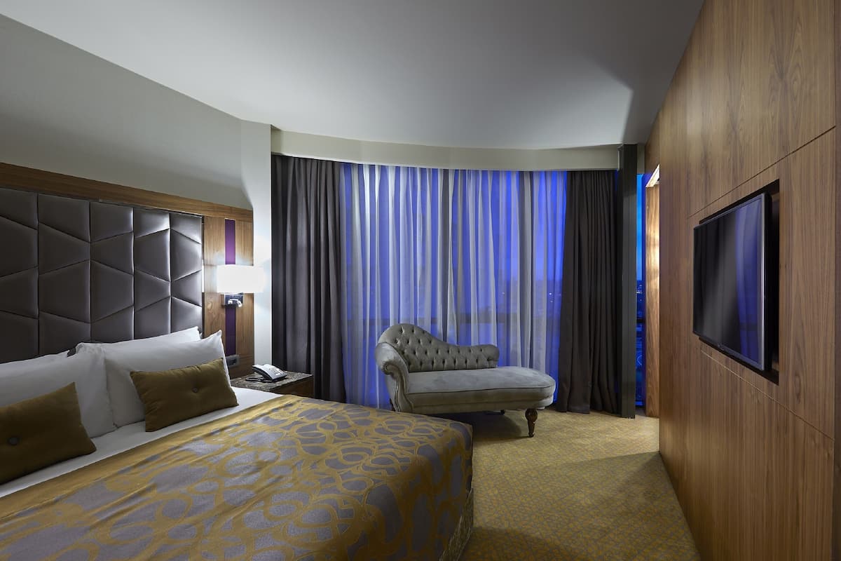 Habitació d'hotel amb un llit gran, un sofà, una televisió i una finestra amb cortines.