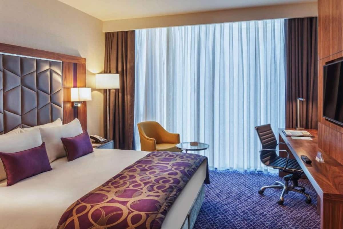 Habitació d'hotel amb un llit gran, una taula de treball, una cadira, un televisor i una finestra amb cortines.