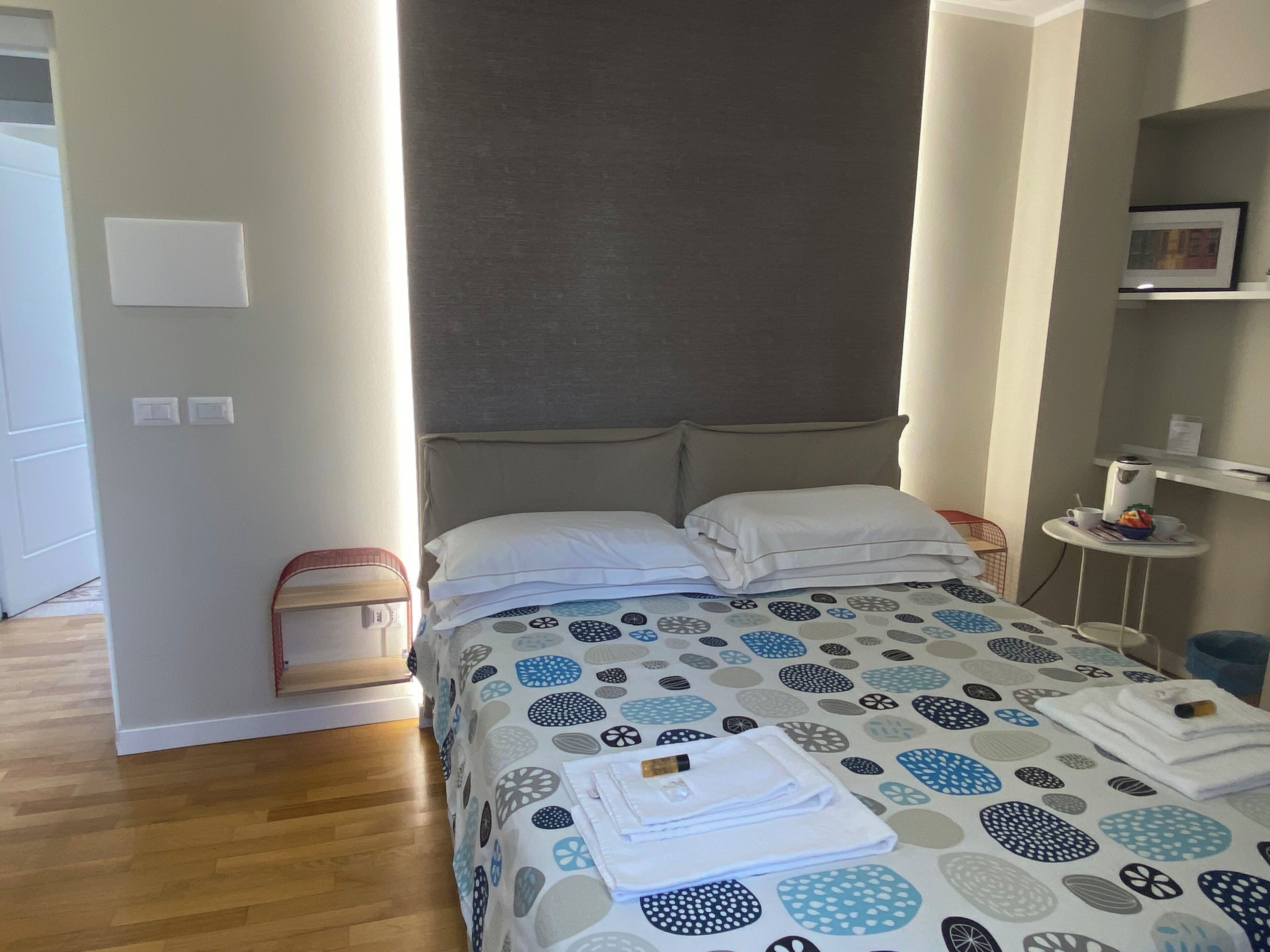 Habitación de hotel con una cama, una mesita de noche pequeña, una silla, un estante con un cuadro y una puerta que da a otra habitación.