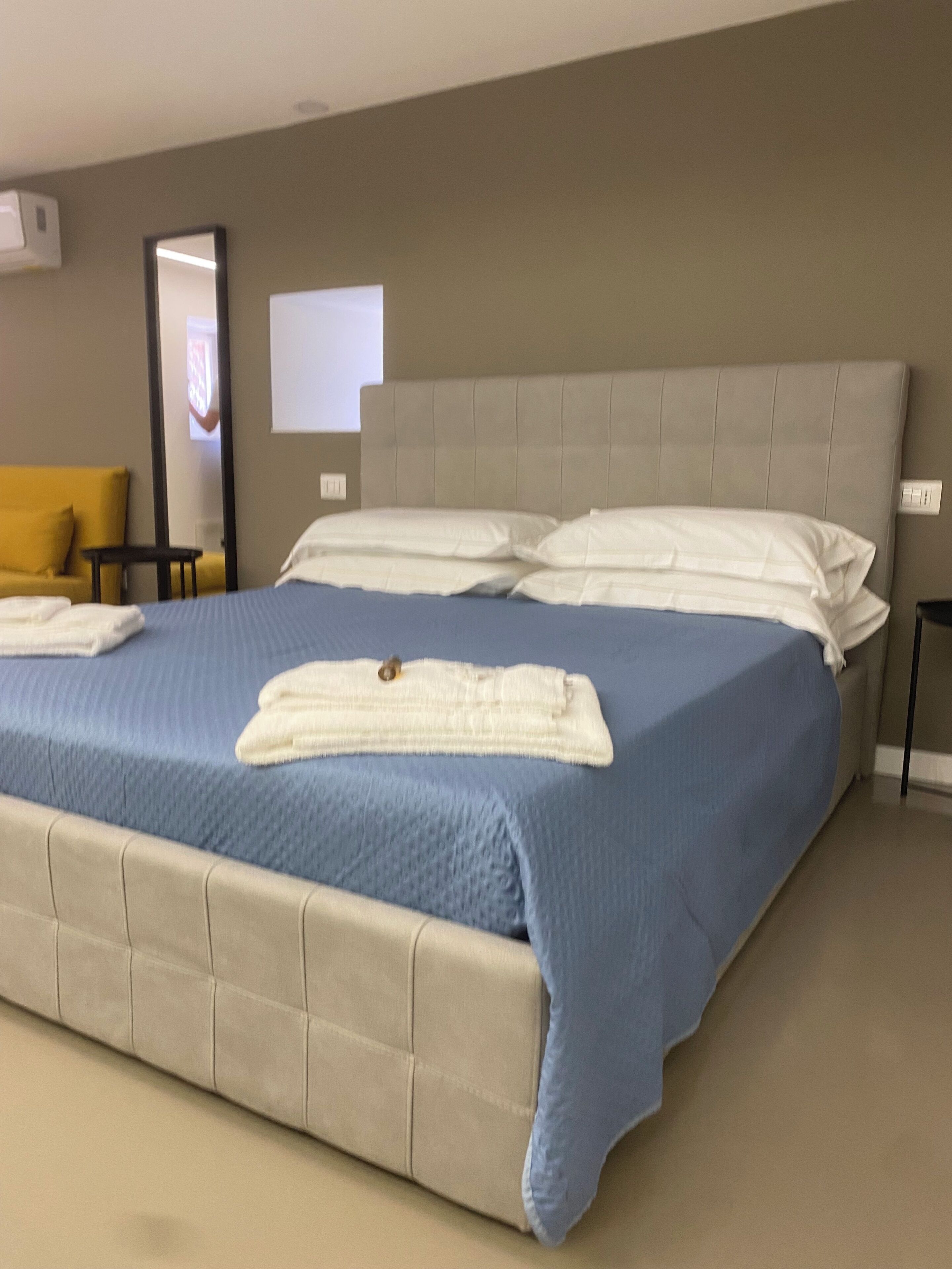 Un lit bien fait, avec une couvre-lit bleue et des oreillers blancs, une tête de lit beige et une petite table de chevet surmontée d’une lampe.