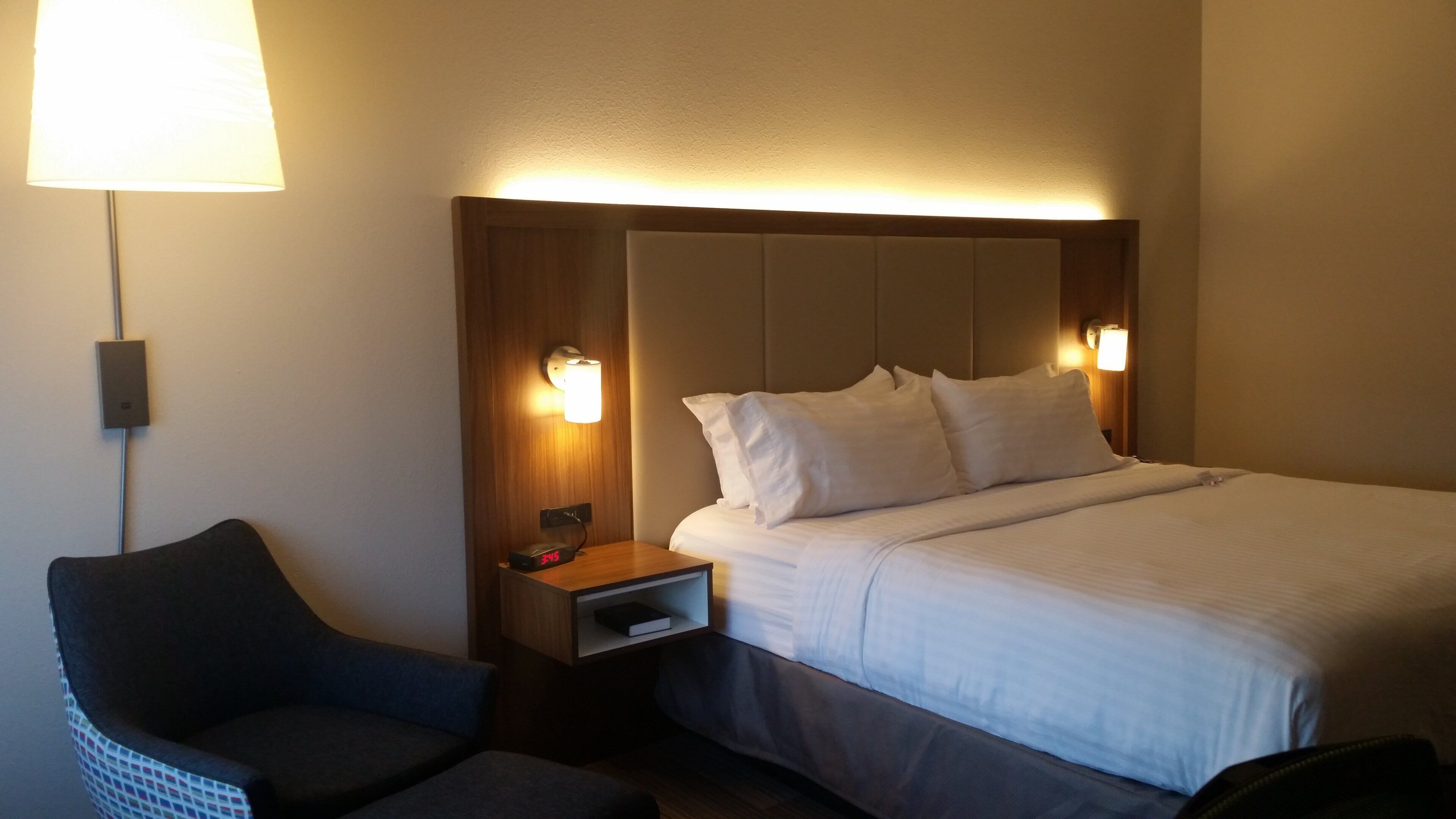A hotel room with a bed, bedside lamps, a nightstand, a chair, and a wall-mounted clock.