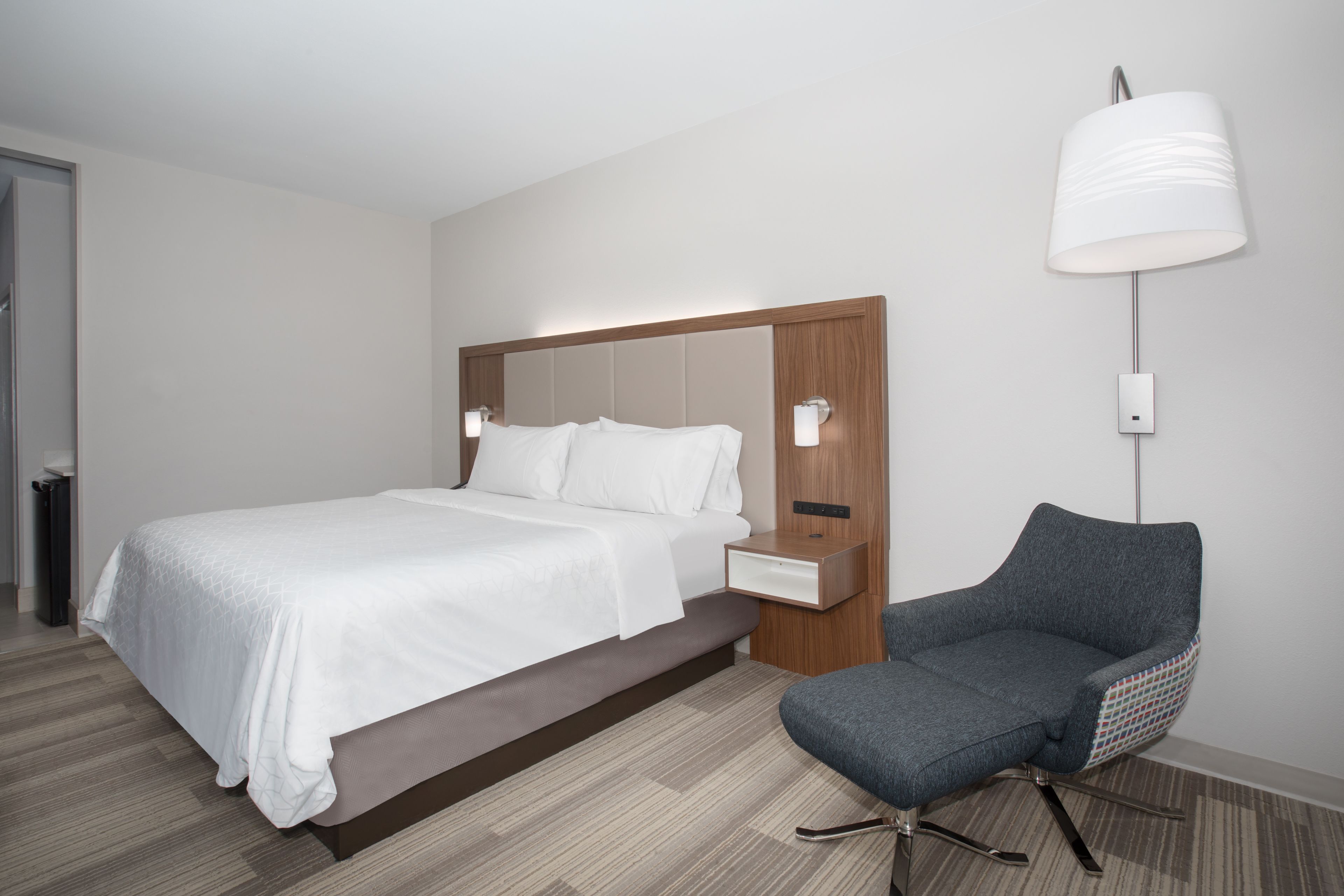 A modern hotel room with a large bed, a nightstand, a chair, and a wall-mounted lamp.