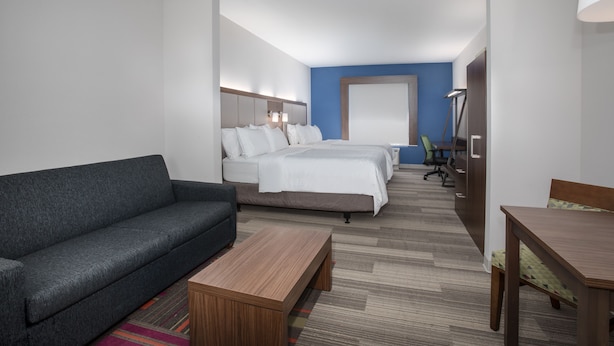 Una habitación de hotel con un sofá gris, una mesa de centro de madera, una cama con ropa de cama blanca, un escritorio con una silla y una lámpara.