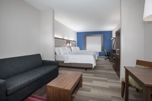 Una habitación de hotel con un sofá gris, una mesa de centro de madera, una cama con ropa de cama blanca, un escritorio con una silla y una lámpara.
