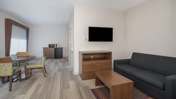A modern hotel room with a sofa, a wooden coffee table, a TV mounted on the wall, a dining area with a table and chairs, and a kitchenette with a microwave and coffee maker.