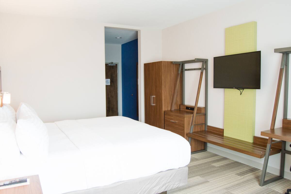 Hotellihuone, jossa on sänky, puinen vaatekaappi, työpöytä tuolilla, seinälle asennettu televisio ja ovi, joka johtaa toiseen huoneeseen.
