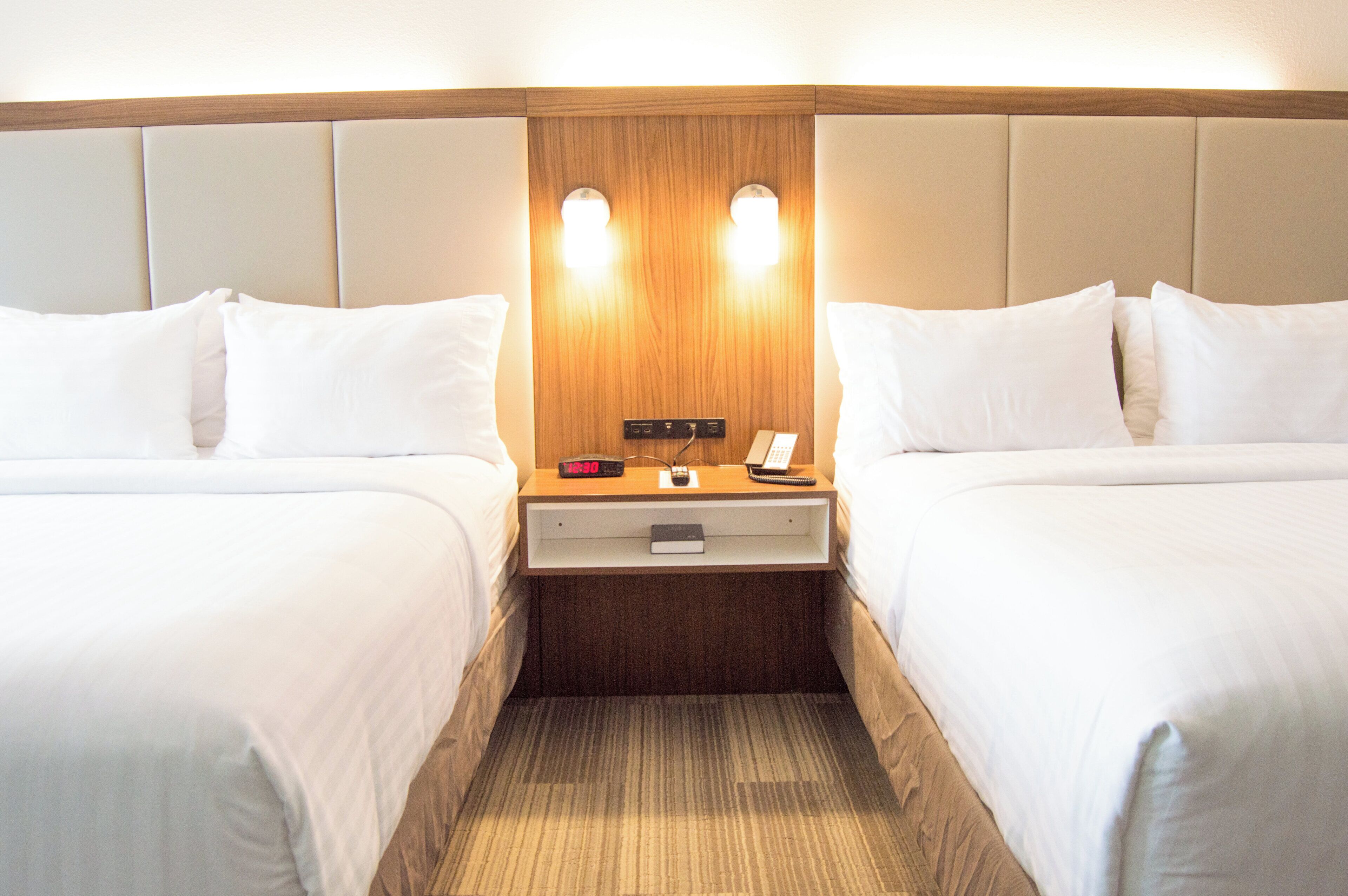 Une chambre d’hôtel avec deux lits, une tête de lit en bois, une table de chevet avec une horloge et un téléphone.