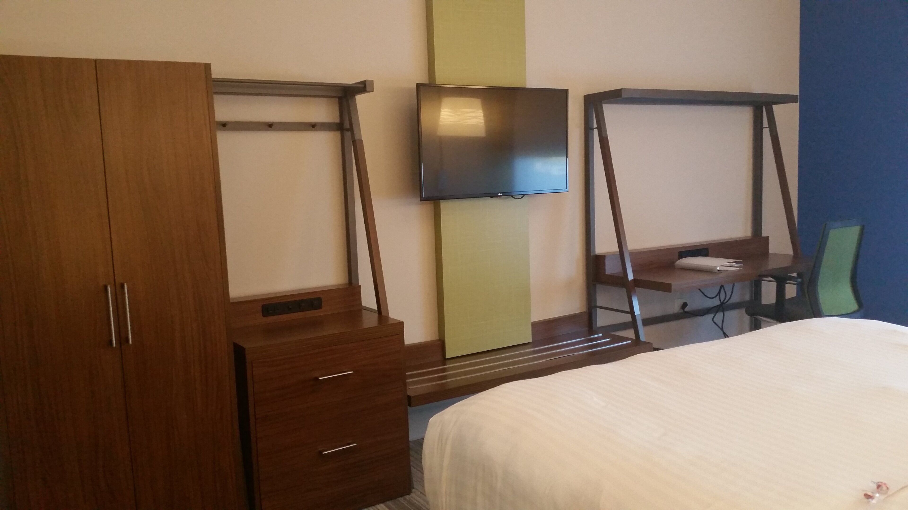 A hotel room with a wooden wardrobe, a television mounted on the wall, a desk with a chair, and a bed with a white bedspread.