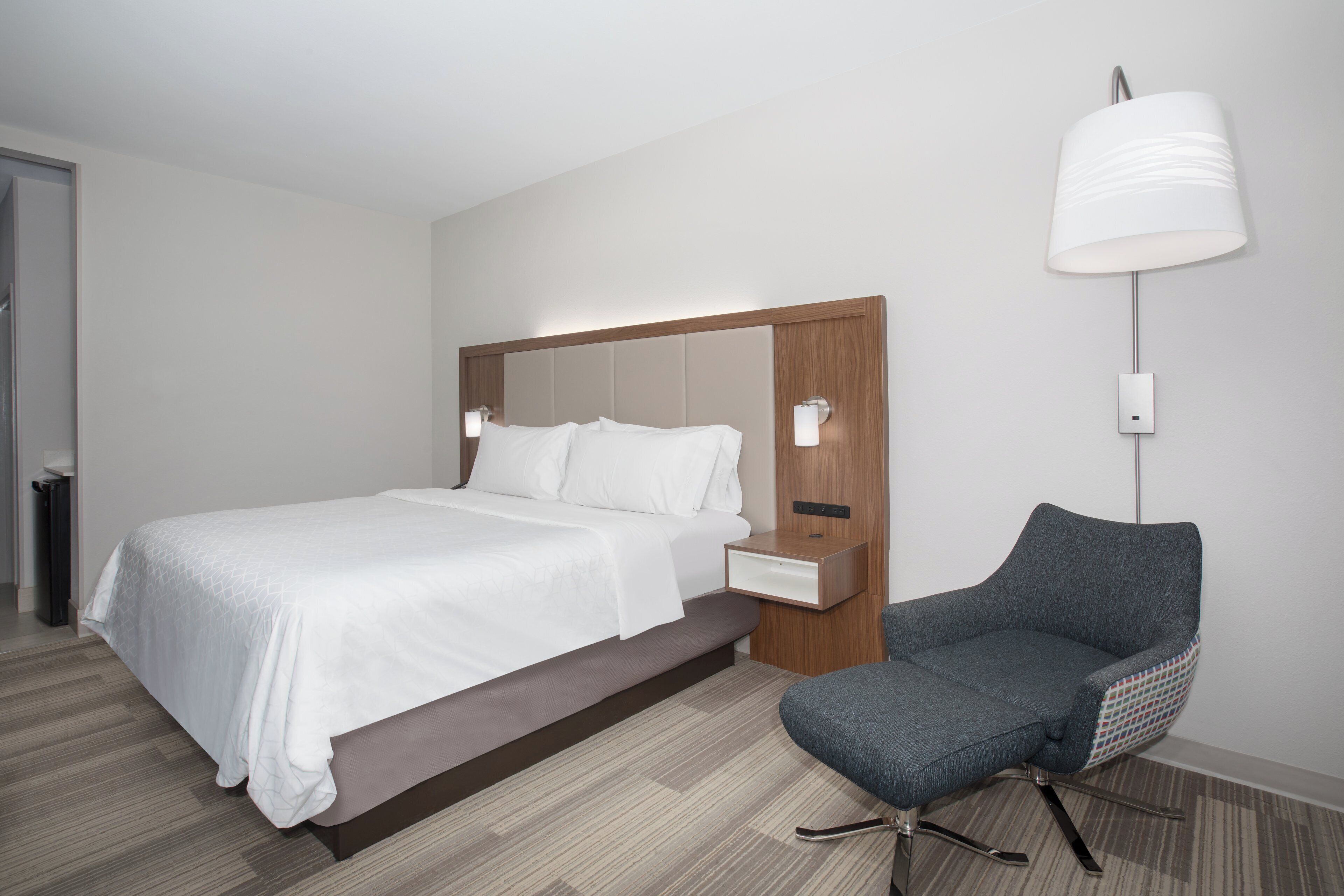 Una habitación de hotel moderna con cama, mesita de noche, lámpara y una silla.
