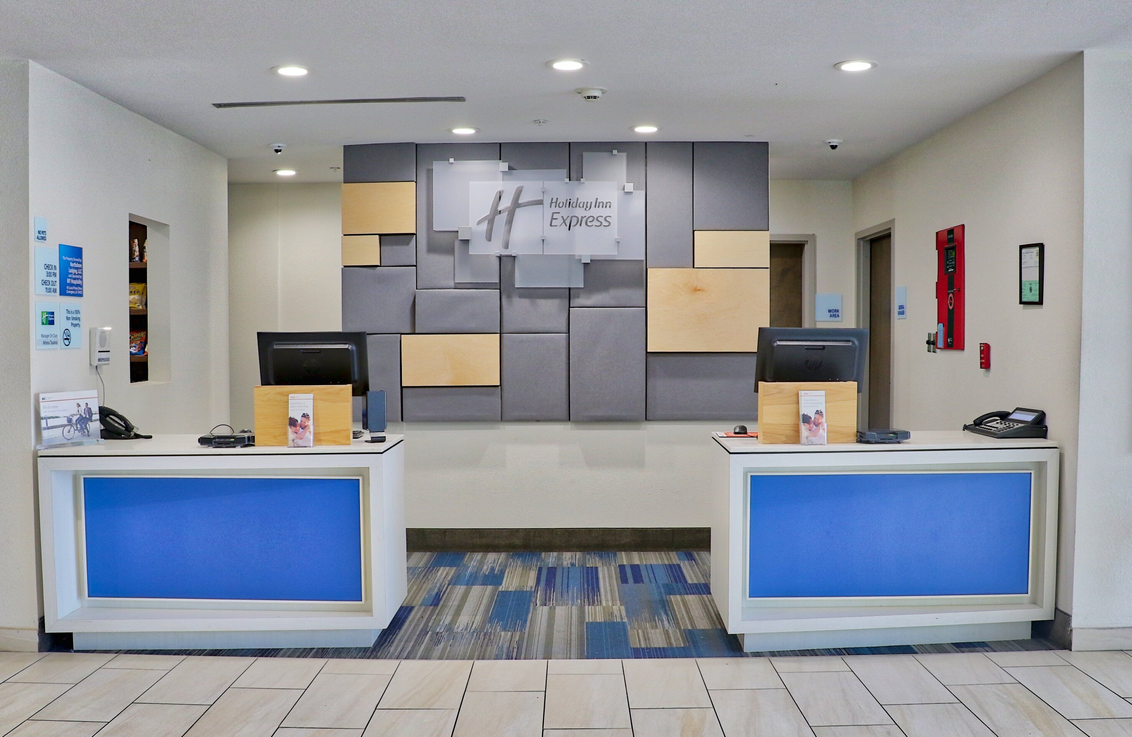 A hotel reception area with two reception desks, a wall-mounted sign for Holiday Inn Express, and a fire alarm on the right.