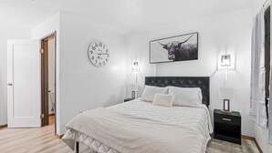 Une chambre à coucher avec un lit, une horloge, un tableau encadré, deux lampes de chevet et une porte.