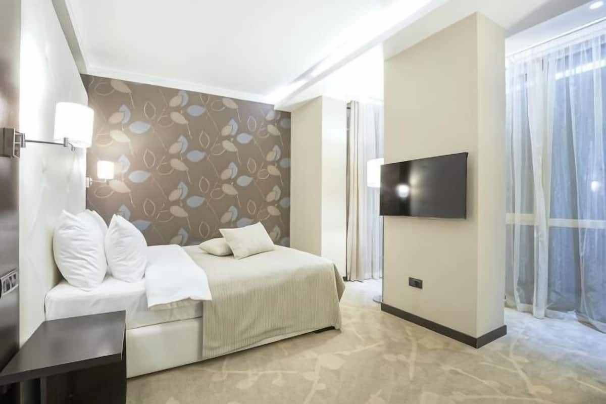A modern hotel room with a bed, a TV, a desk, and a large window.