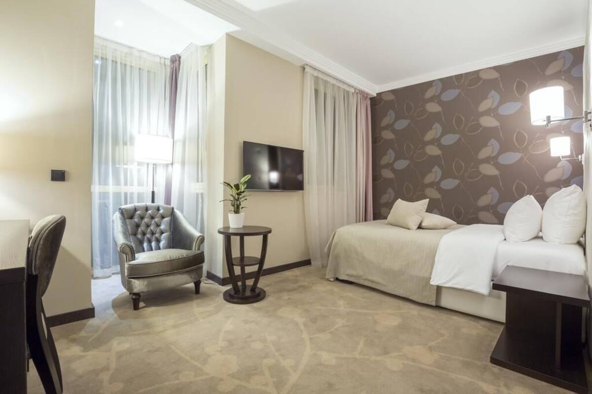 A hotel room with a bed, a chair, a small table with a plant, a TV on the wall, and a patterned wall.