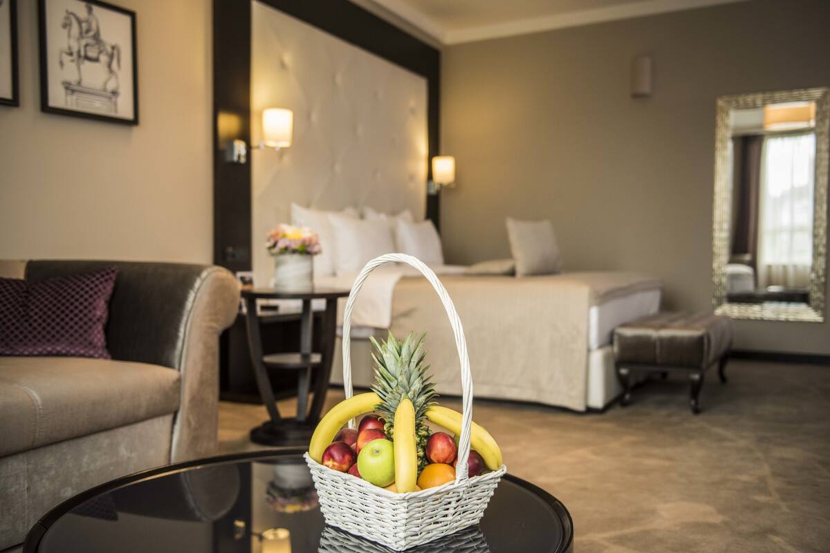A hotel room with a bed, a dining table, a sofa, and a fruit basket on a glass table.