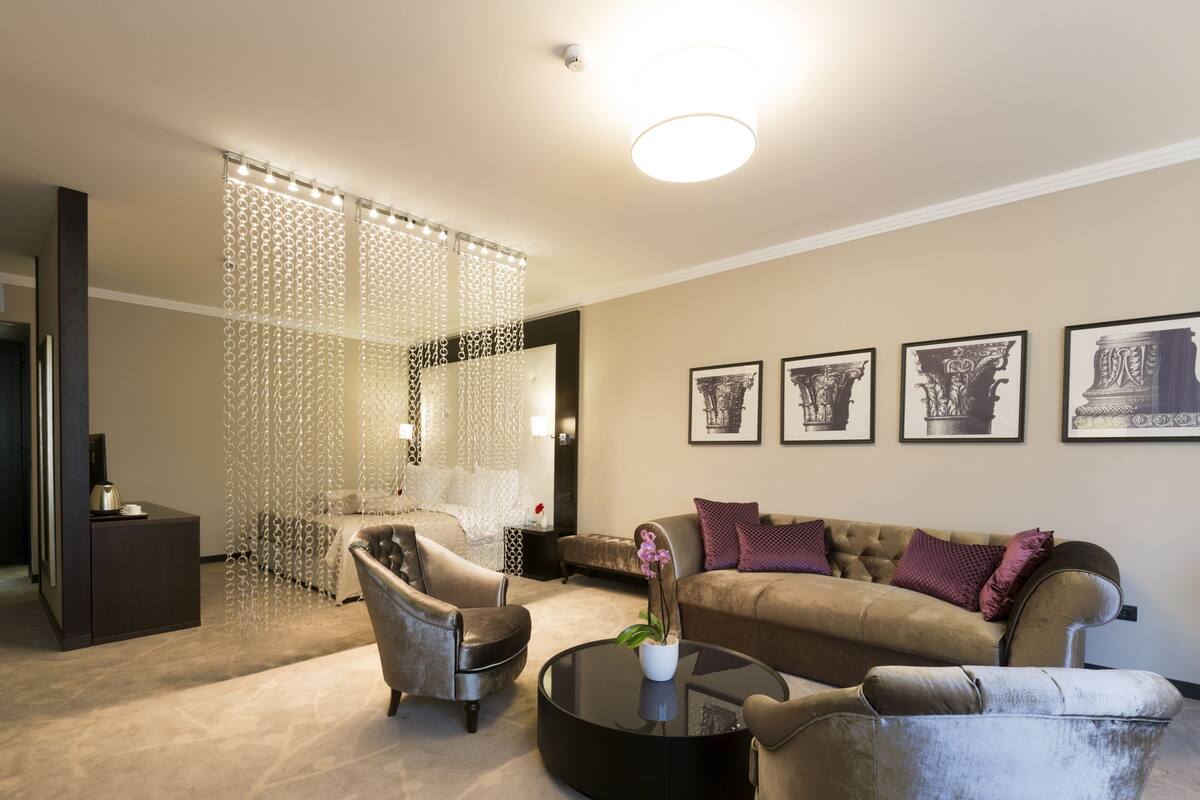 A modern living room with a sofa, armchair, and coffee table. There are framed pictures on the wall and a decorative curtain.