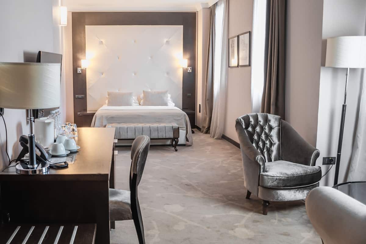 A hotel room with a large bed, a desk with a lamp, a chair, and a bedside table with a phone and glasses.