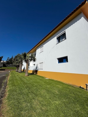 A white building with a yellow base, a red roof, and a well-maintained lawn with palm trees. - Hotel J.S.F. (Horta)