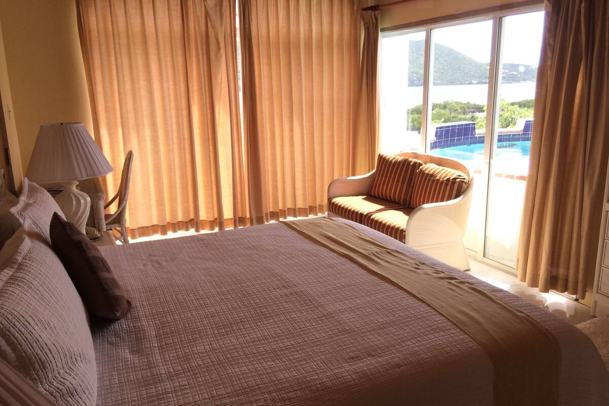 Quarto de hotel com uma cama grande, uma cadeira, um abajur e vista para uma piscina.