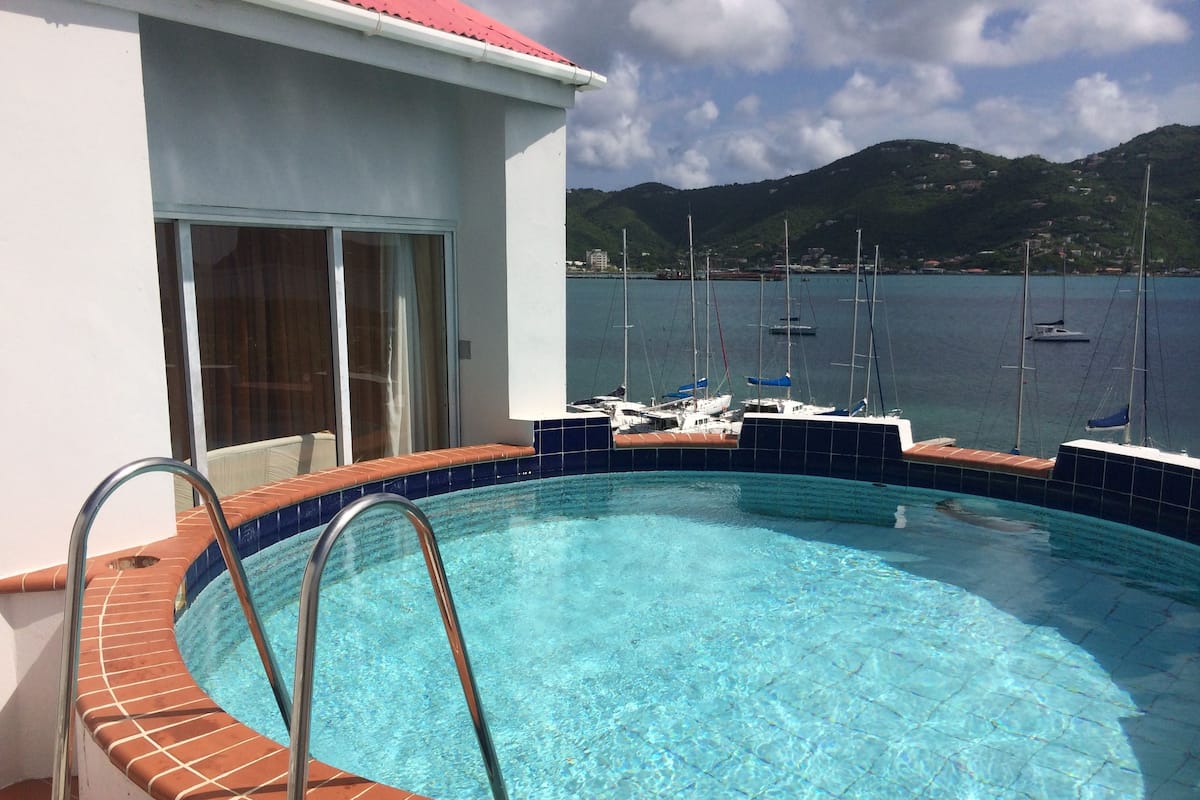 Uma piscina com água azul cristalina, rodeada por um deck revestido em azulejos. Ao fundo, barcos na água e uma paisagem montanhosa.