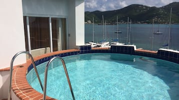 A swimming pool with a clear blue water, surrounded by a tiled deck. In the background, there are boats on the water and a mountainous landscape.