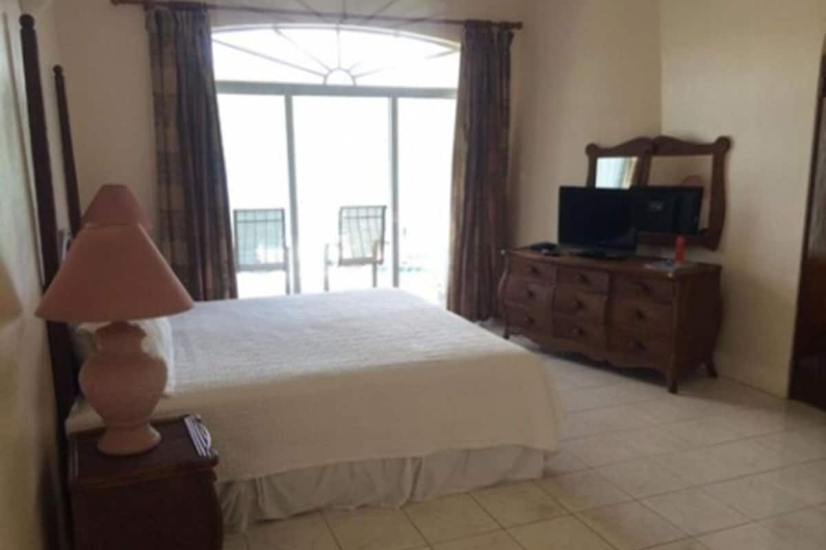 Quarto com cama, cômoda, luminária, televisão e varanda com cortinas.
