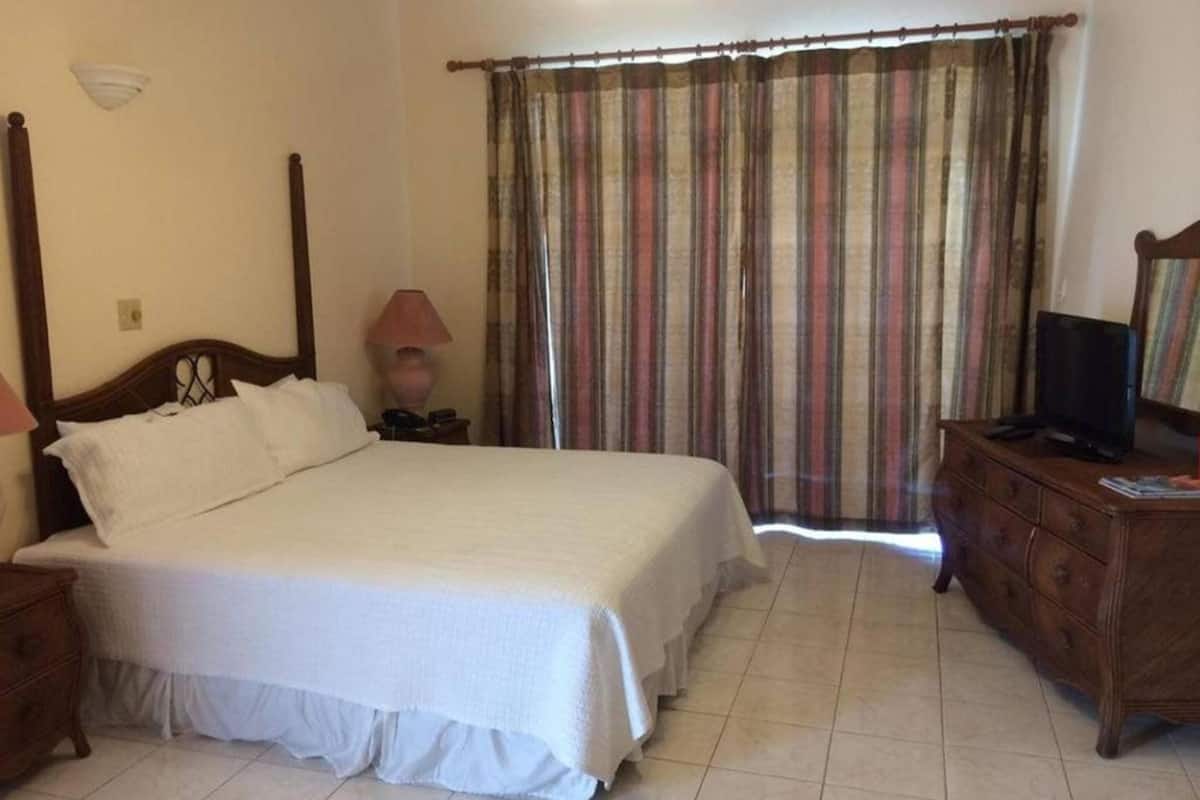 Quarto com uma cama grande, penteadeira de madeira, mesa de cabeceira com abajur e televisão sobre a penteadeira.