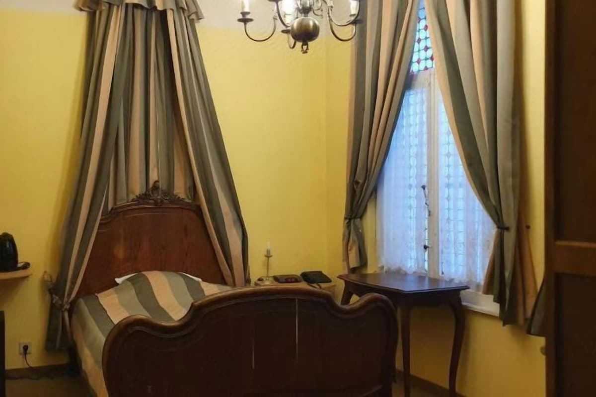 A bedroom with a bed, a bedside table, a window with curtains, and a chandelier.