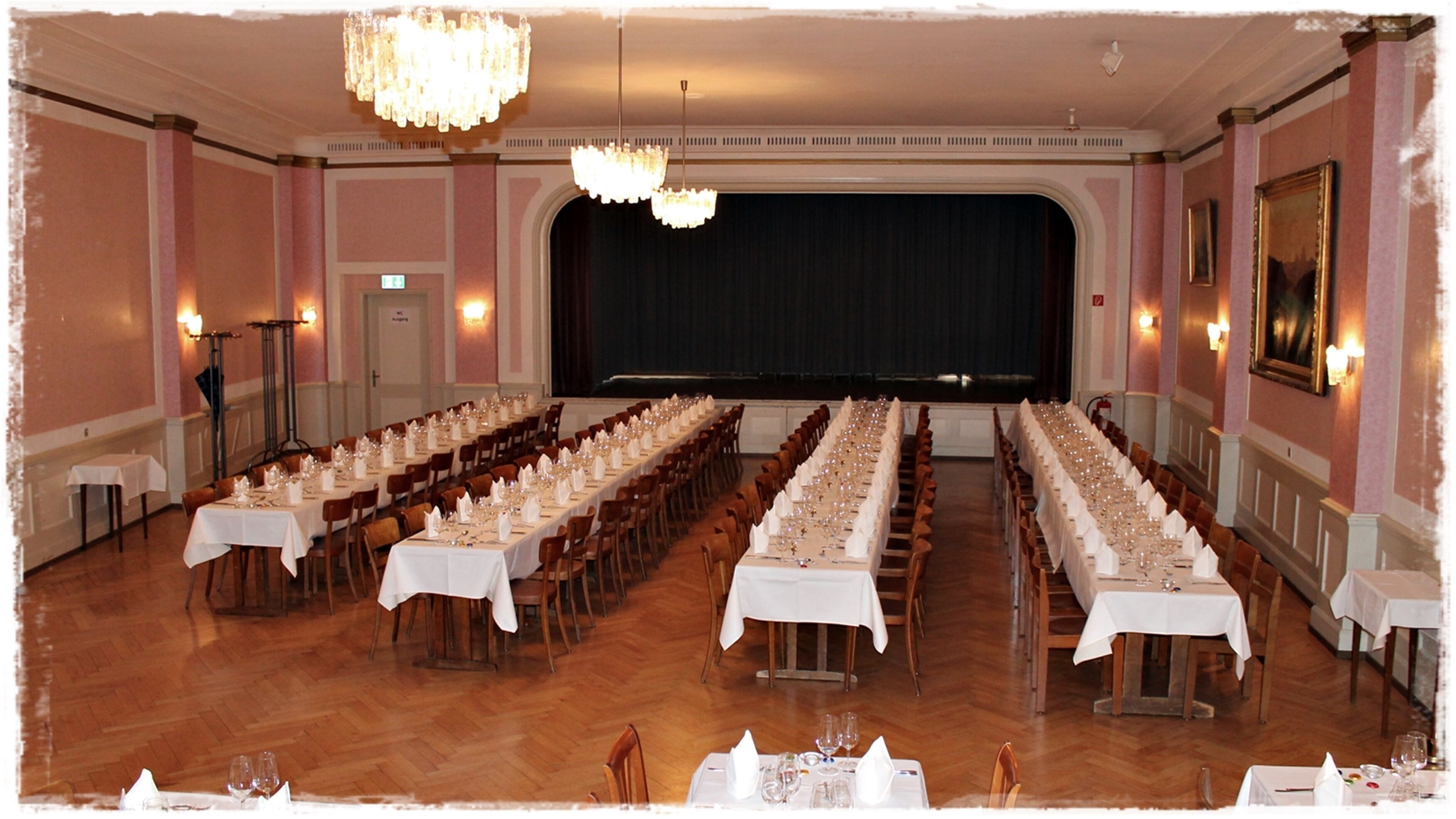 A banquet hall with long tables set for a formal event, chandeliers, and a stage with a curtain backdrop.