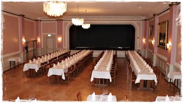 A banquet hall with long tables set for a formal event, chandeliers, and a stage with a curtain backdrop.