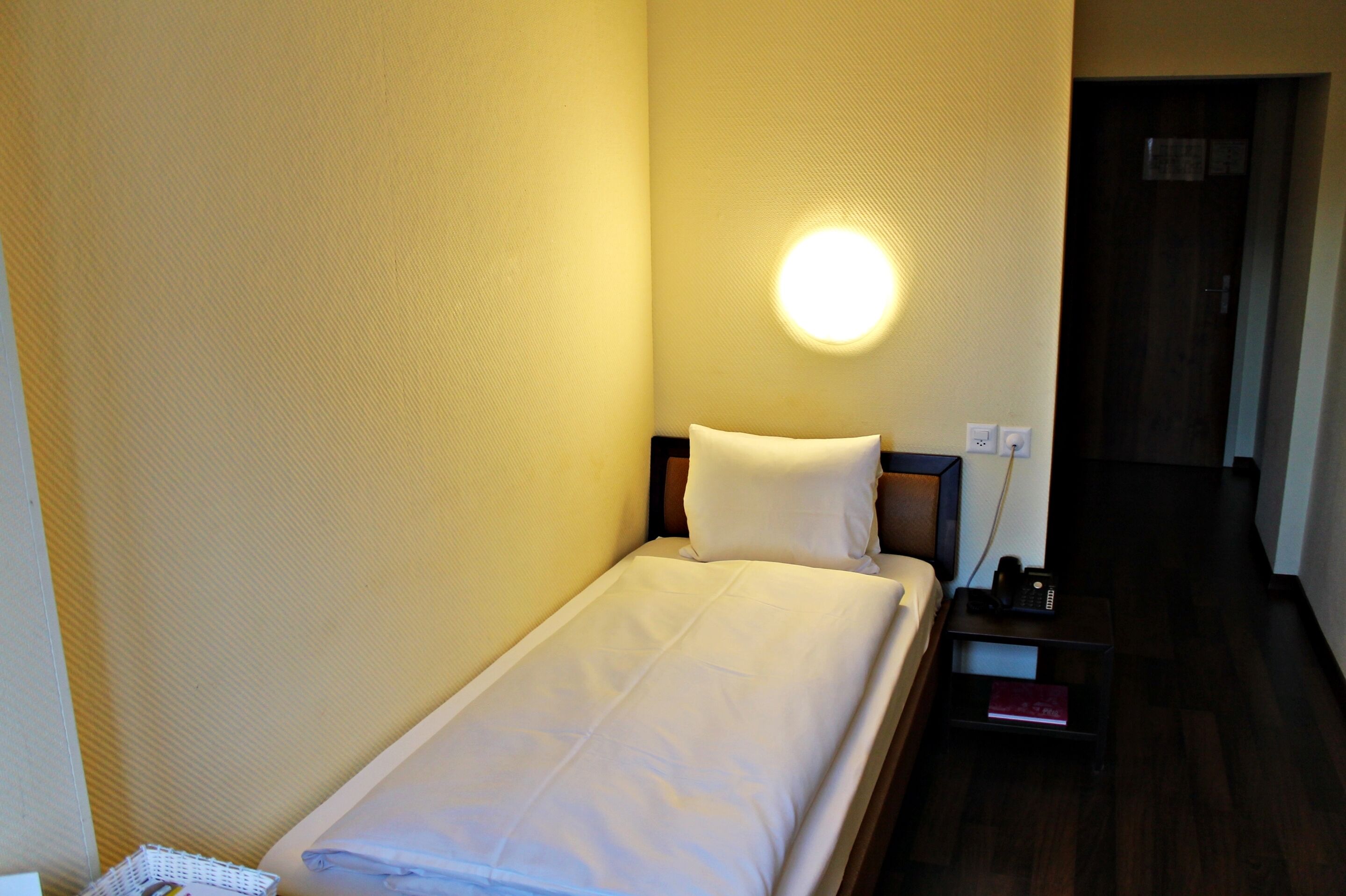 Een klein, keurig opgemaakt bed met witte lakens in een compacte kamer, met een nachtkastje, een wandlamp en een deur naar een andere kamer.