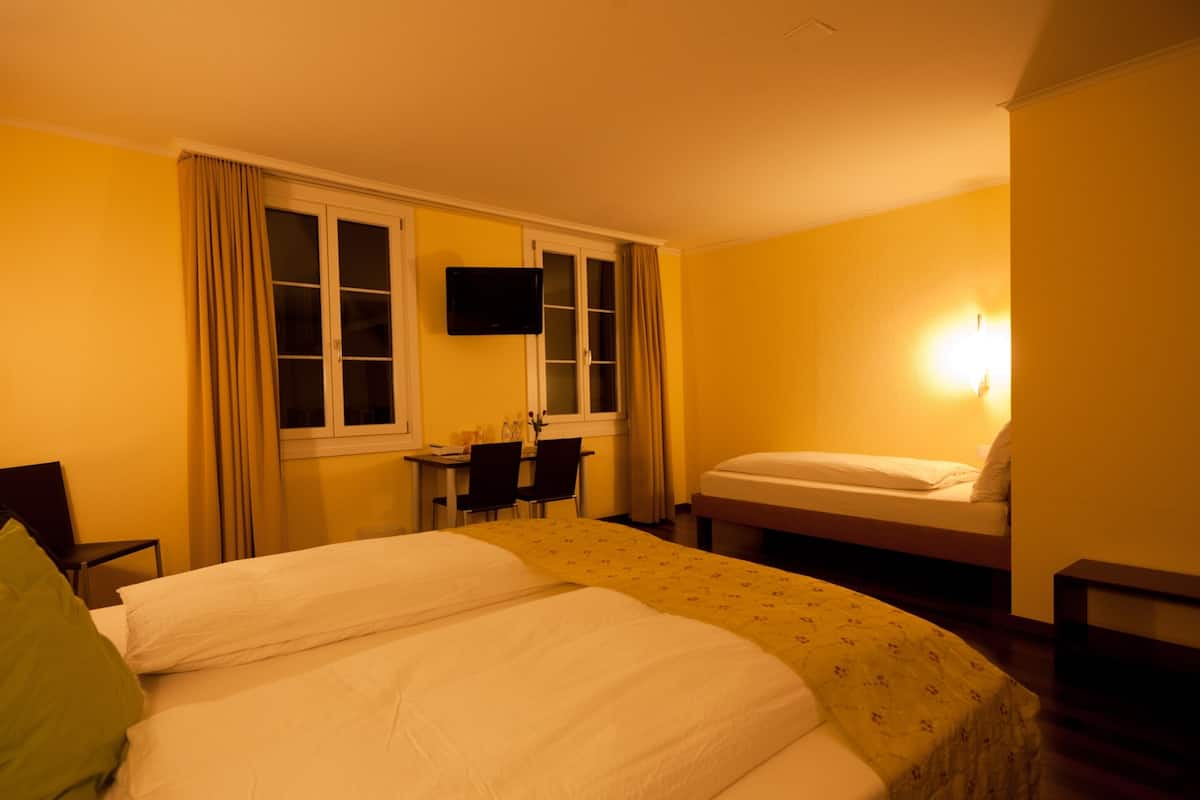 A hotel room with two beds, a desk with chairs, a TV, and a window with curtains.