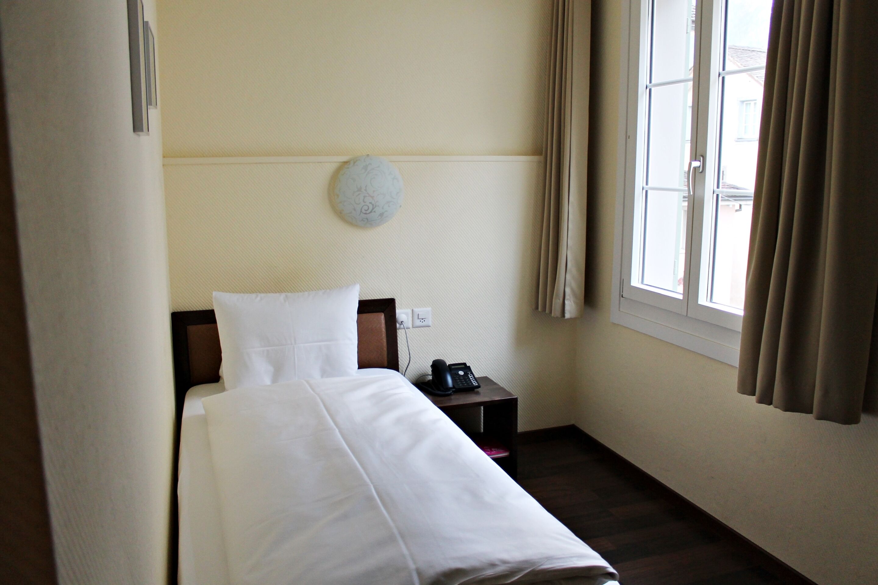 Une chambre d’hôtel avec un lit, une table de chevet avec un téléphone, une lampe fixée au mur et une fenêtre avec des rideaux.