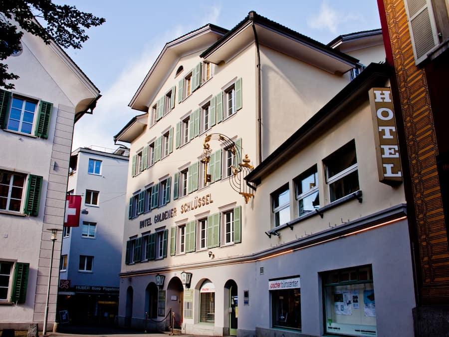 A hotel with a classic European facade, featuring a central entrance with a staircase, multiple windows with green shutters, and a sign indicating it is a hotel.